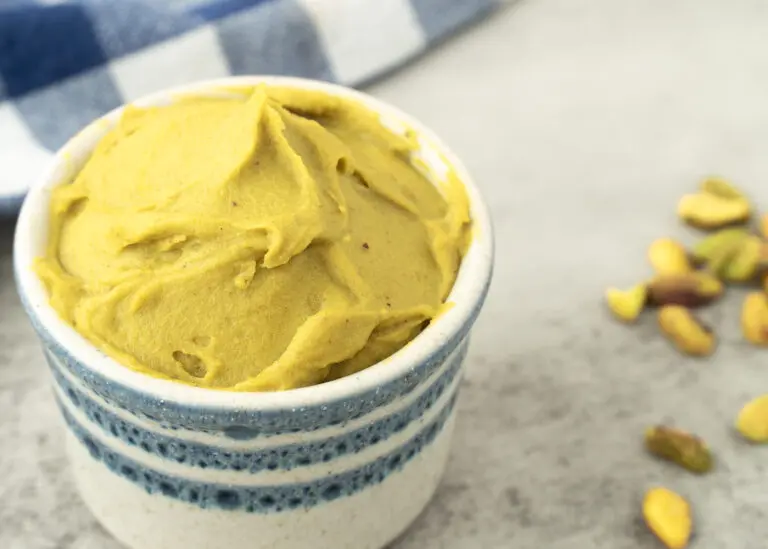 Pistachio Paste in a white ramekin with blue stripes. Pistachio nuts are pictured on the right side and a blue and white checked napkin are pictured in the left corner.