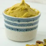 white ramekin with blue stripes filled with pistachio paste and raw pistachio nuts on the side.