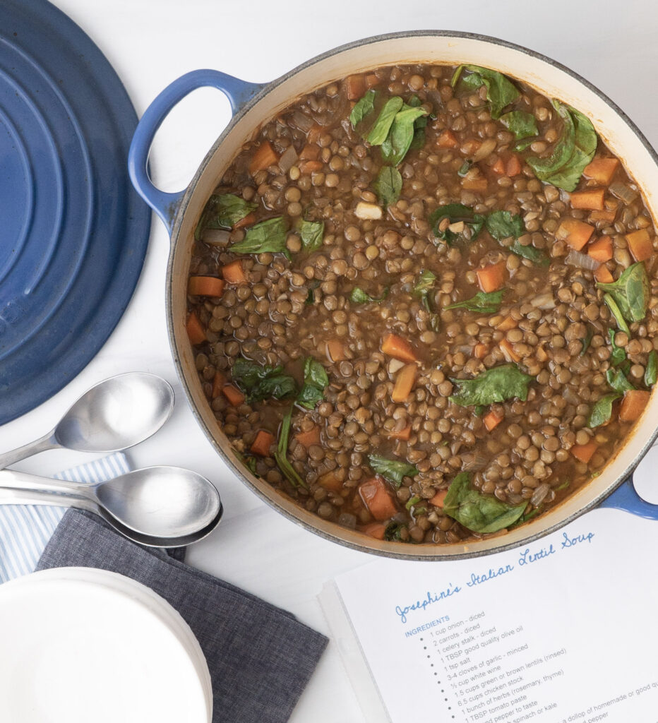 Overhead photo of a pot of lentil soup with spoons showing and a blue pot lid and and blue and white striped napkin.