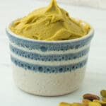 white ramekin with blue stripes filled with pistachio paste and raw pistachio nuts on the side.