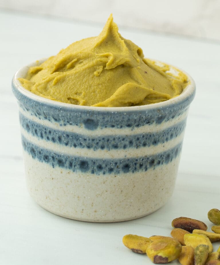 white ramekin with blue stripes filled with pistachio paste and raw pistachio nuts on the side.