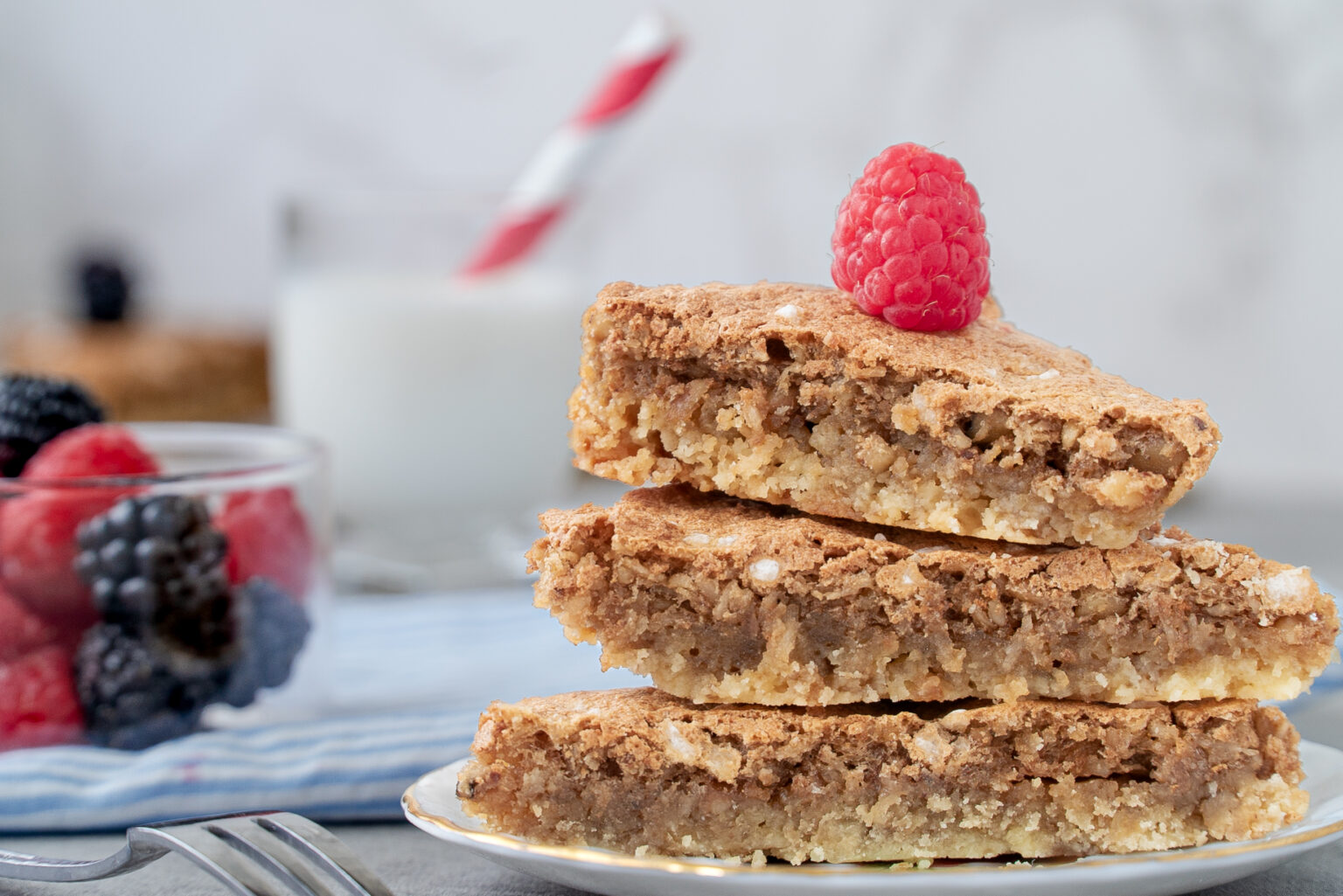 Butternut Bars | stack of bars on a plate with a raspberry on top. Berries on the side and a glass of milk with a red and white striped straw in the background