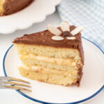 Slice of vanilla cake with chocolate frosting, almond flower decoration, and a fork on the plate.