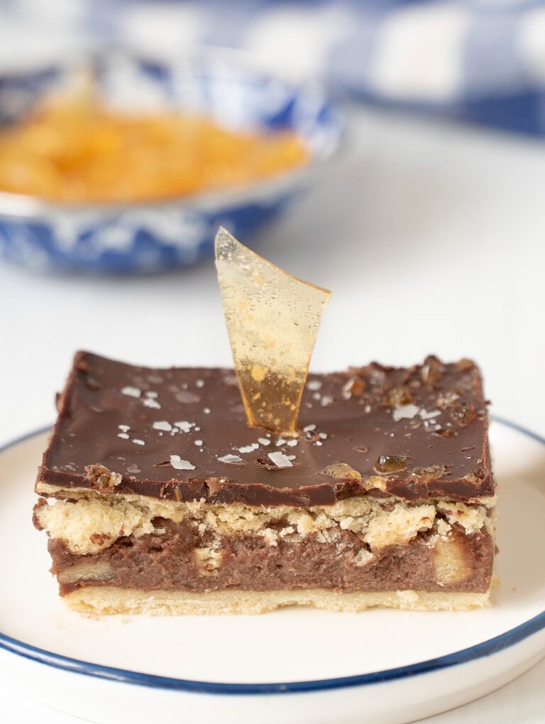 Slice of Kati Tészta (Hungarian Chocolate Rum Torte) with burnt caramel shards and a sprinkle of sea salt, blurred blue and white bowls in the background