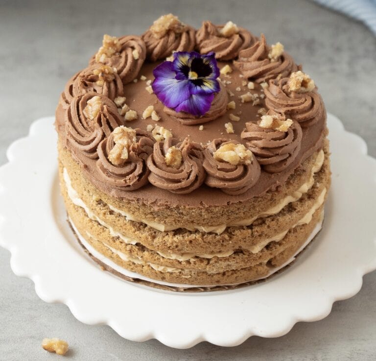Close up photo of a hungarian nut torte with mocha frosting, candied walnuts and decorated wth a fresh purple pansy.