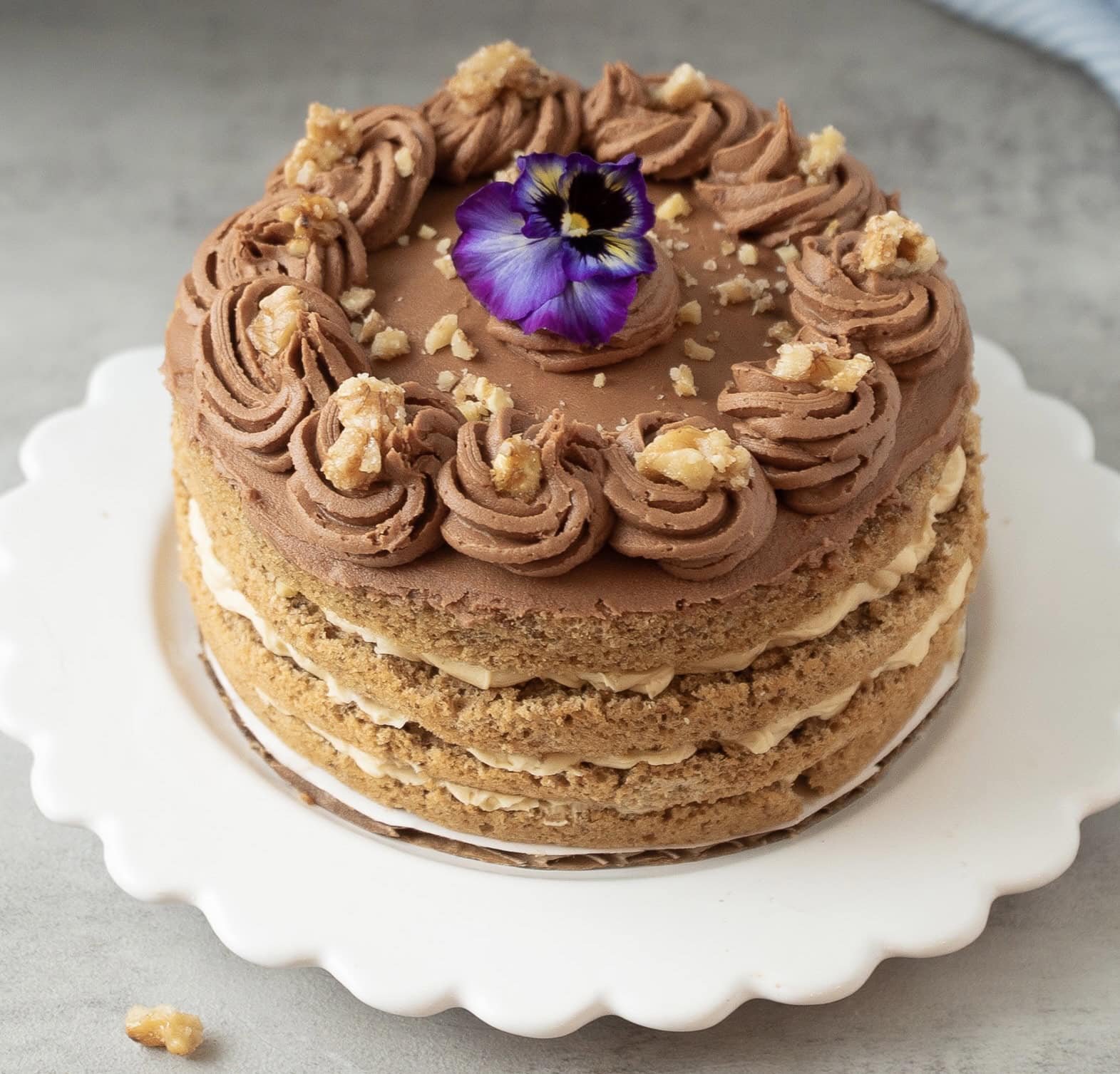 Close up photo of a hungarian nut torte with mocha frosting, candied walnuts and decorated wth a fresh purple pansy.