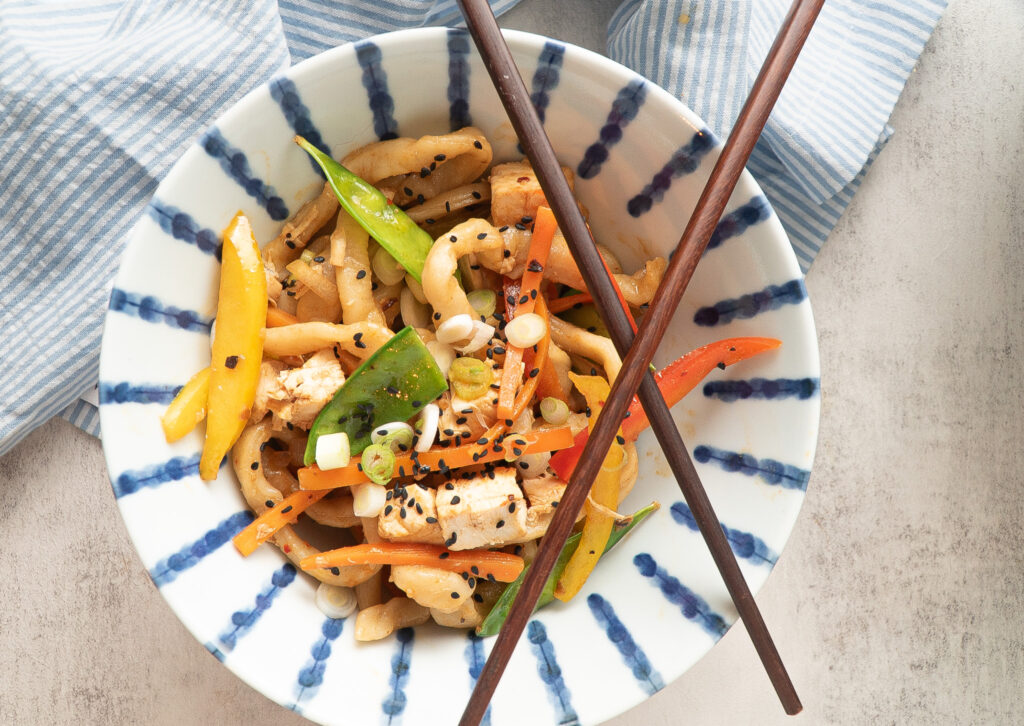 Colorful weeknight udon stir fry in a blue and white striped bowl, topped with crossed chopsticks and served with a light blue napkin.