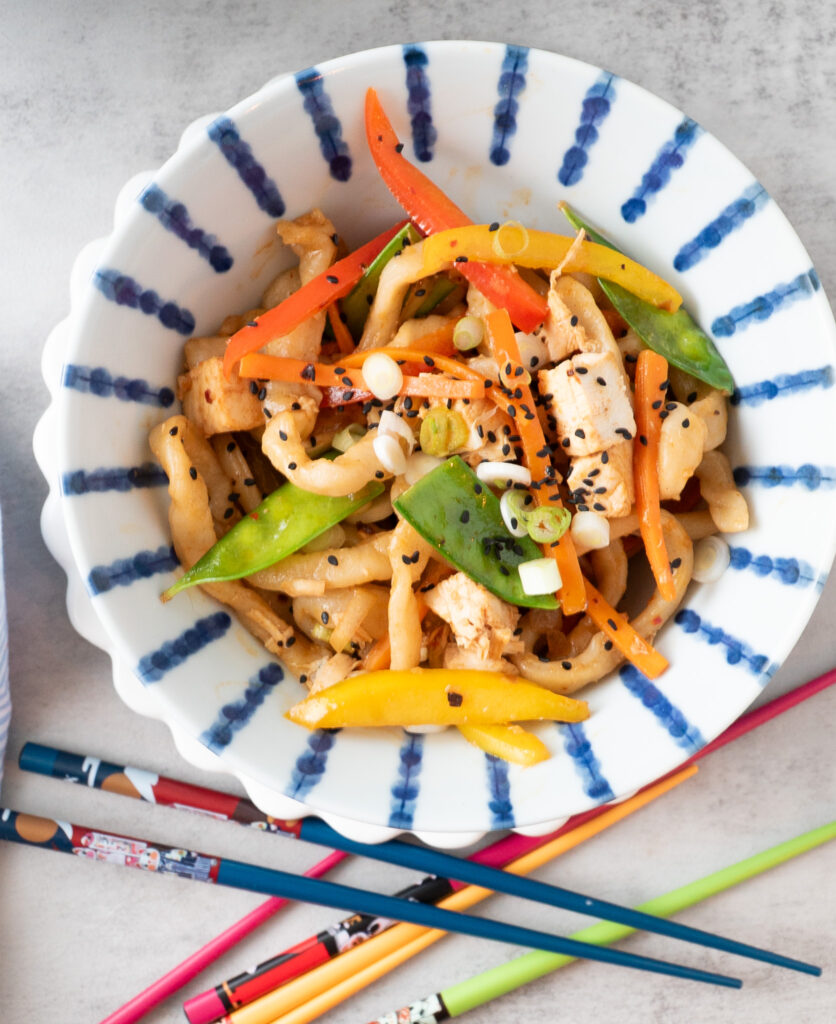 A colorful bowl of vegetables, chicken and fresh udon noodles with colorful chopsticks laying beside the bowl