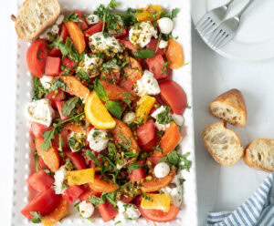 Colorful Summer Sunset Caprese Salad served on a large white rectangular plate with watermelon, oranges, tomatoes, mozzarella, burrata, herbs, and pistach