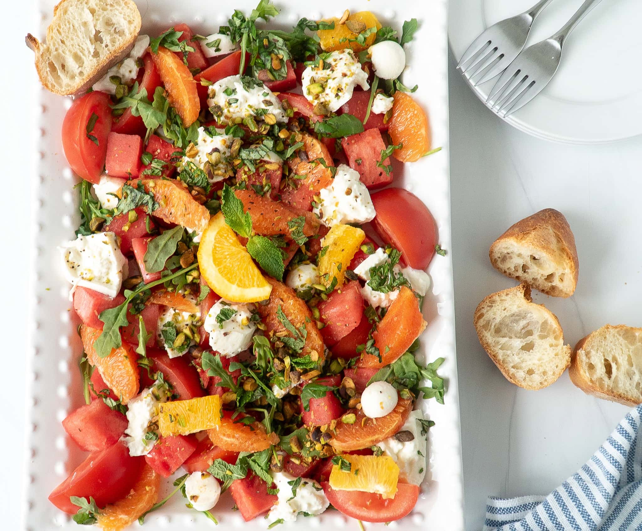 Colorful Summer Sunset Caprese Salad served on a large white rectangular plate with watermelon, oranges, tomatoes, mozzarella, burrata, herbs, and pistach