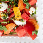 Close-up of Summer Sunset Caprese Salad with watermelon, oranges, tomatoes, burrata, and herbs on a beaded white plate