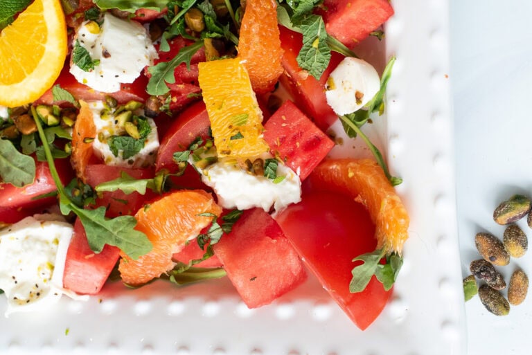 Close-up of Summer Sunset Caprese Salad with watermelon, oranges, tomatoes, burrata, and herbs on a beaded white plate