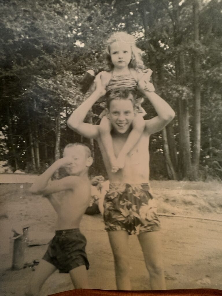 Black and white vintage photo of three siblings at the lake — little sister sitting on big brother’s shoulders, little brother flexing his muscle