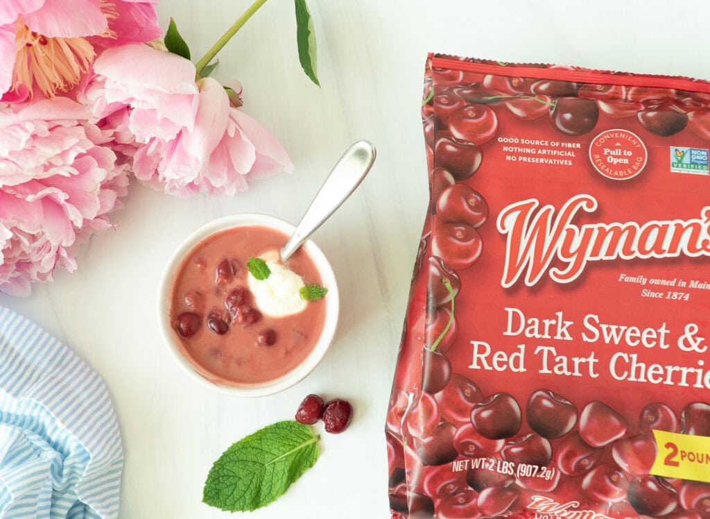 Overhead shot of chilled Hungarian sour cherry soup garnished with cream and herbs. Flowers  displayed next to a bag of Wyman’s frozen cherry blend.