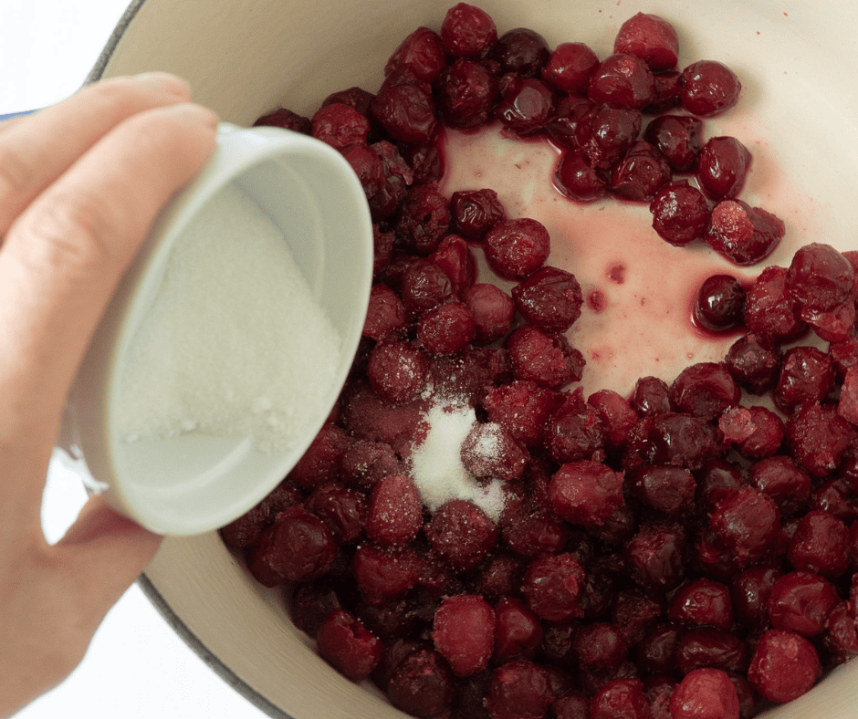Close-up of granulated sugar being poured into a pot of thawing sour cherries on the stove.