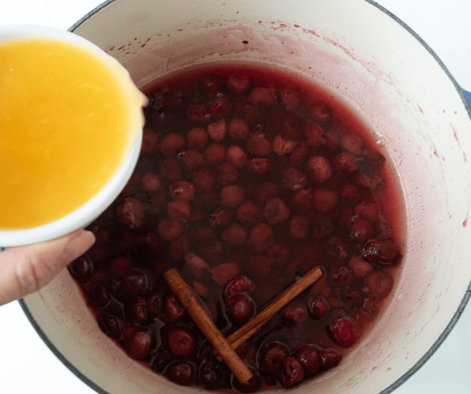 Fresh orange juice being poured into the pot of simmering sour cherries and spices.