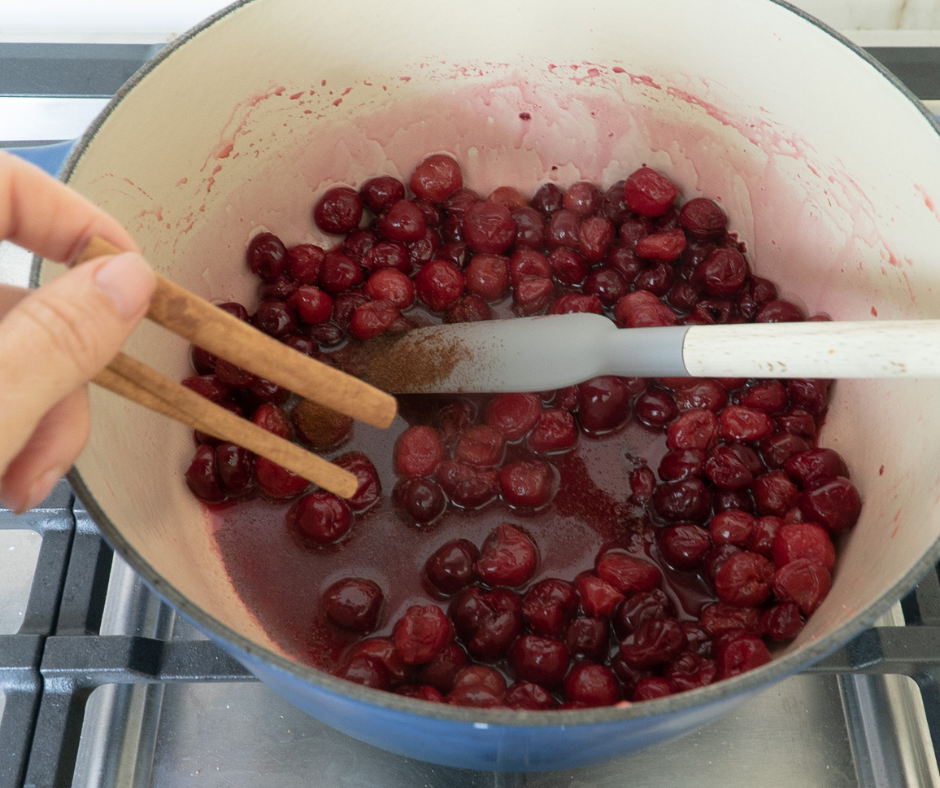 Cinnamon sticks being added to simmering sour cherries in a large pot