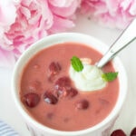 Bowl of chilled Hungarian sour cherry soup garnished with a swirl of cream and fresh herbs, with soft-focus flowers in the background