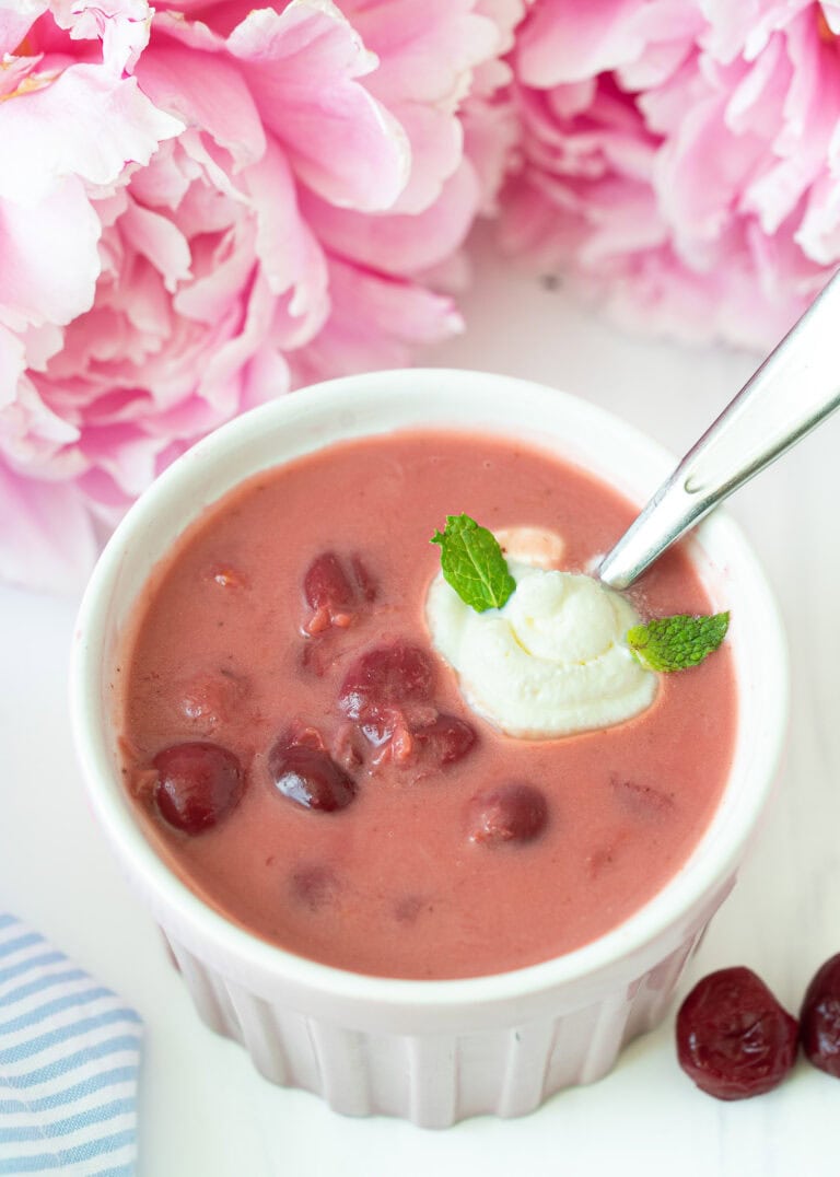 Bowl of chilled Hungarian sour cherry soup garnished with a swirl of cream and fresh herbs, with soft-focus flowers in the background