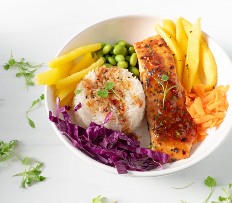 Overhead shot of a Thai salmon rice bowl topped with cilantro sprigs, colorful vegetables, mango, and avocado, served with lime wedges.