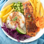 Close-up of a Thai salmon rice bowl showing salmon, colorful vegetables, mango, and avocado, served with a fork on a blue napkin.