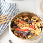 Close-up of a Tex-Mex Chicken Quinoa Bowl with colorful veggies, black beans, quinoa, and shredded chicken, styled with a striped napkin and bamboo fork.