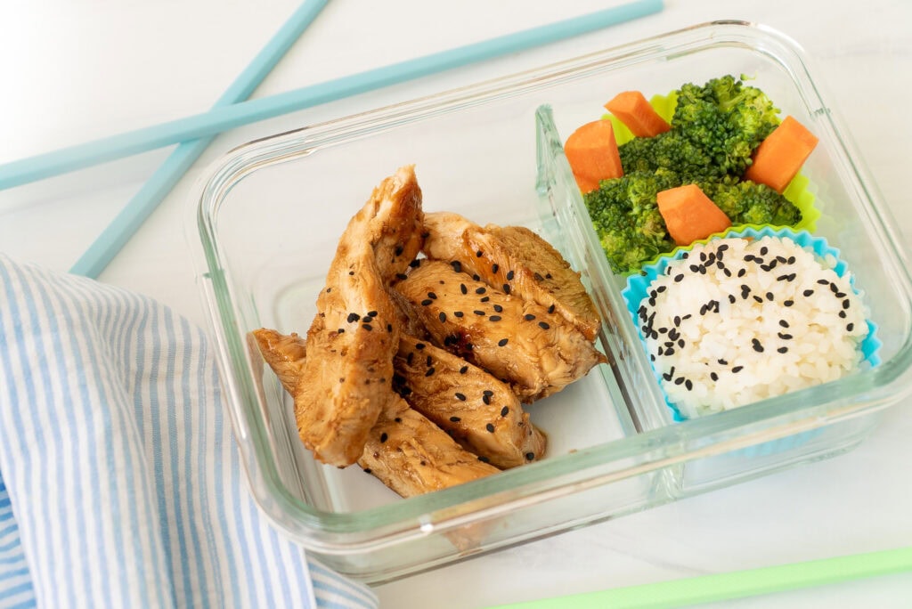 Top view of Teriyaki Chicken Bento Box with rice and broccoli, placed on a blue-and-white striped napkin with blue and green chopsticks