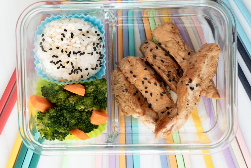 Teriyaki Chicken Bento Box with rice and steamed broccoli, served with rainbow-colored chopsticks on a light background
