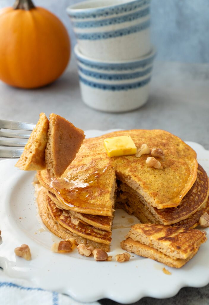 A stack of pumpkin spice protein pancakes with a first bite on a fork pictured. In the background is a peek at a mini pumpkin and a blue and white striped ramekin.
