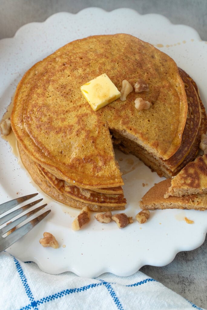 Close Up Photo of Pumpkin Spice Protein Pancakes with a bite out of them and drizzled with maple syrup, walnuts and topped with a pat of butter.