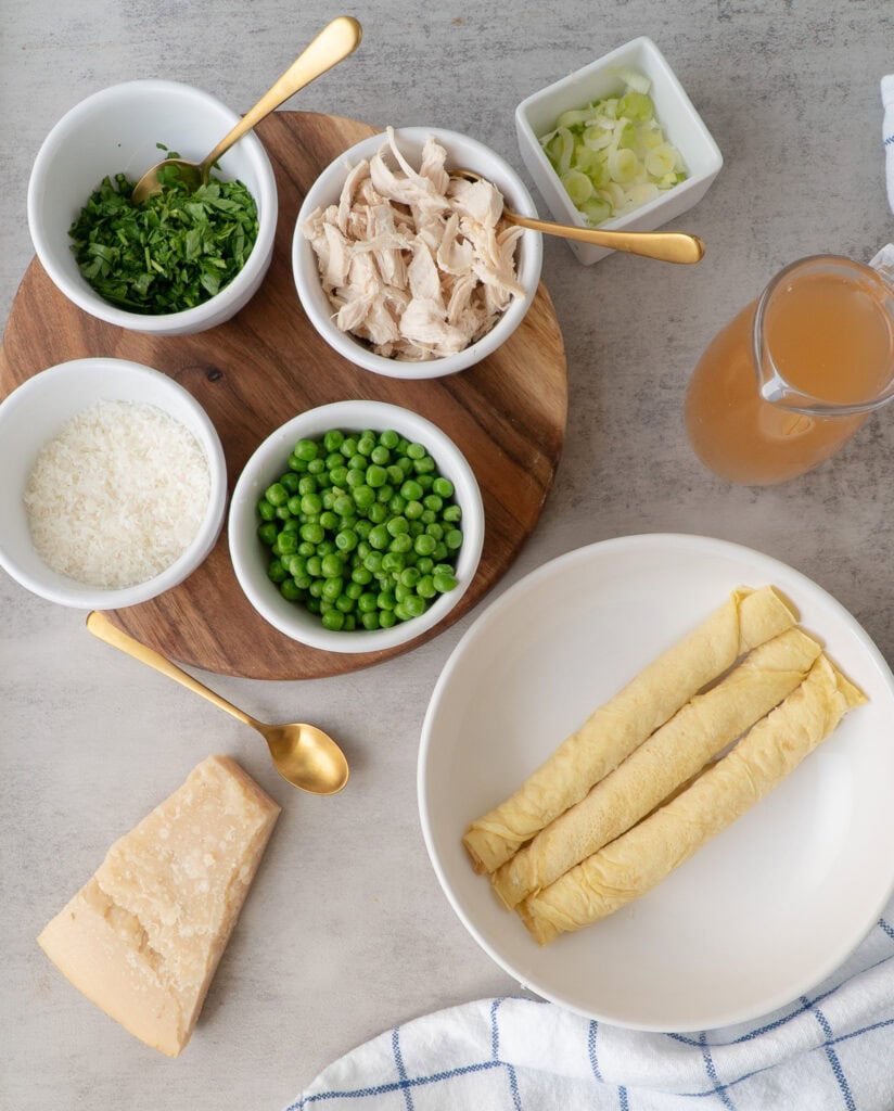  three scrippelle in a bowl with a pitcher of broth and a side board with ramekins filled with toppings - chicken, peas, parmesan and parsley.