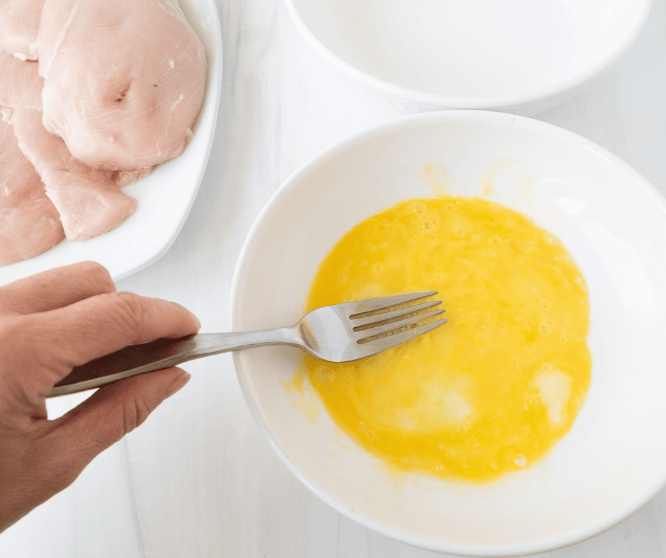 hand using a fork to mix an egg in a bowl for dipping chicken before breading 