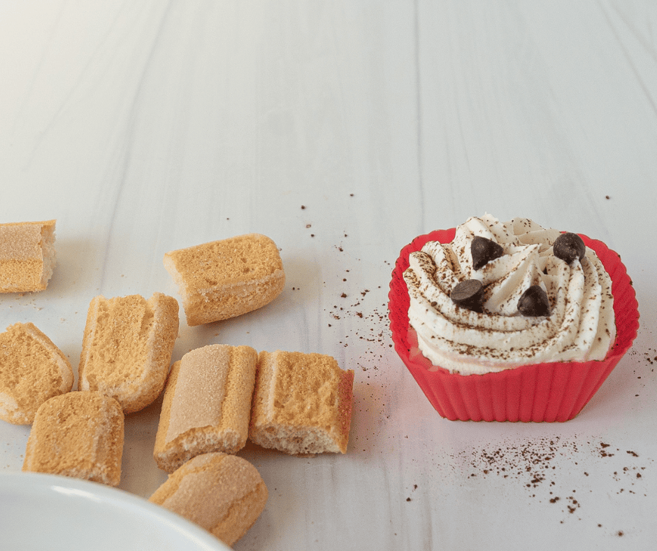 Finished tiramisu cup next to lady finger cookies and a dusting of cocoa
