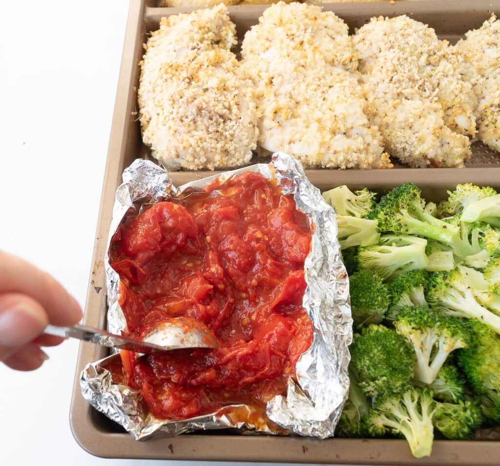 Hand crushing tomatoes with a spoon on a sheet pan while making easy sheet pan chicken parmesan