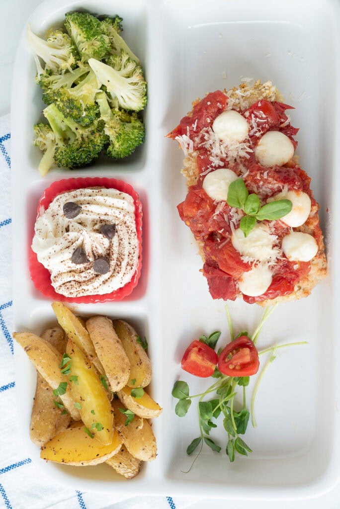 Easy Sheet Pan Chicken Parmesan Dinner Recipe Aerial Shot | A rectangular plate with chicken and sauce with cheese on it, broccoli, roasted potatoes and a dessert cube with a fork visible.