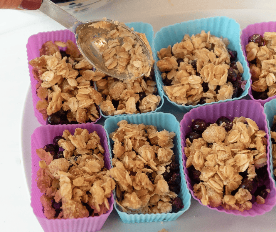 PROCESS picture of a spoon adding crumb topping on top of the blueberry filling in silicone cups