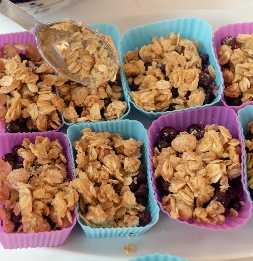Wild blueberry crisps in blue and purple colored silicone cups with a spoon adding ingredients to a cup. 