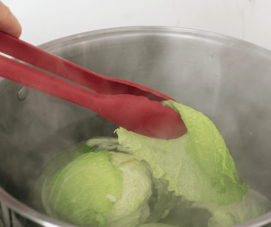 Red tongs pulling individual cabbage leaves out of a pot of boiling water.