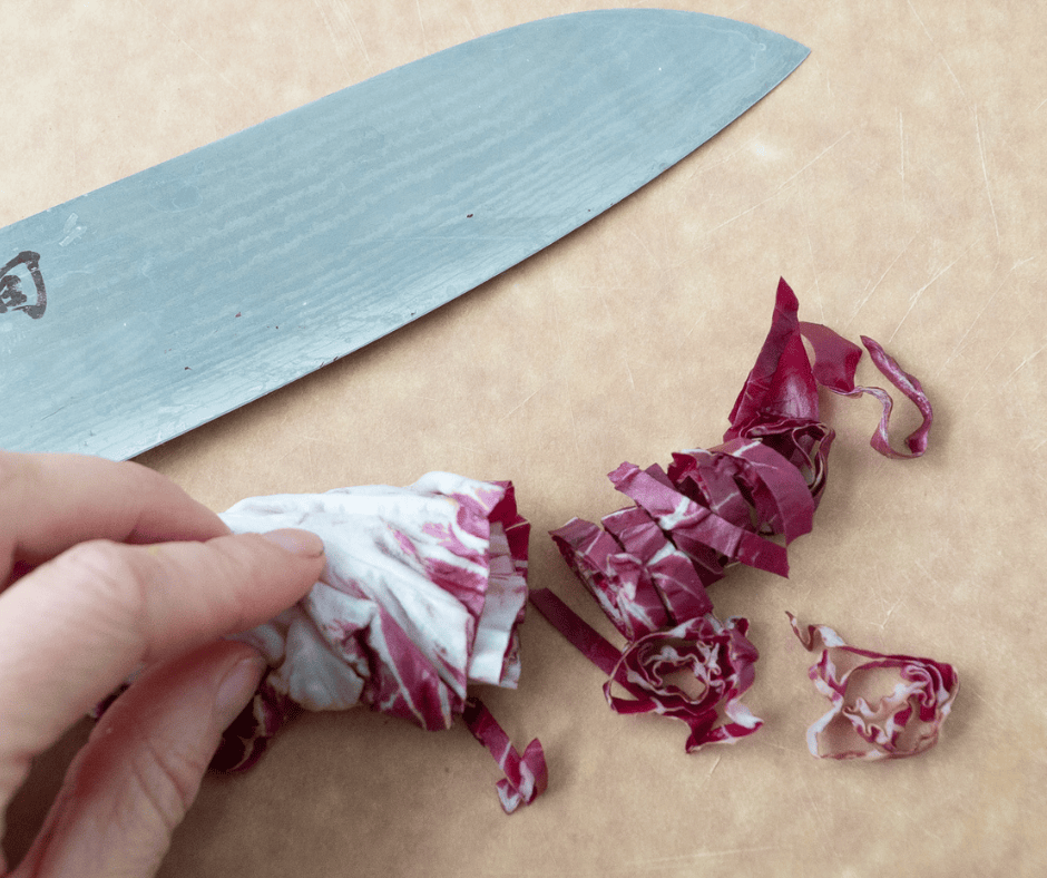 A hand, a knife and a leaf of red radicchio lettuce rolled and sliced horizontally into ribbons.