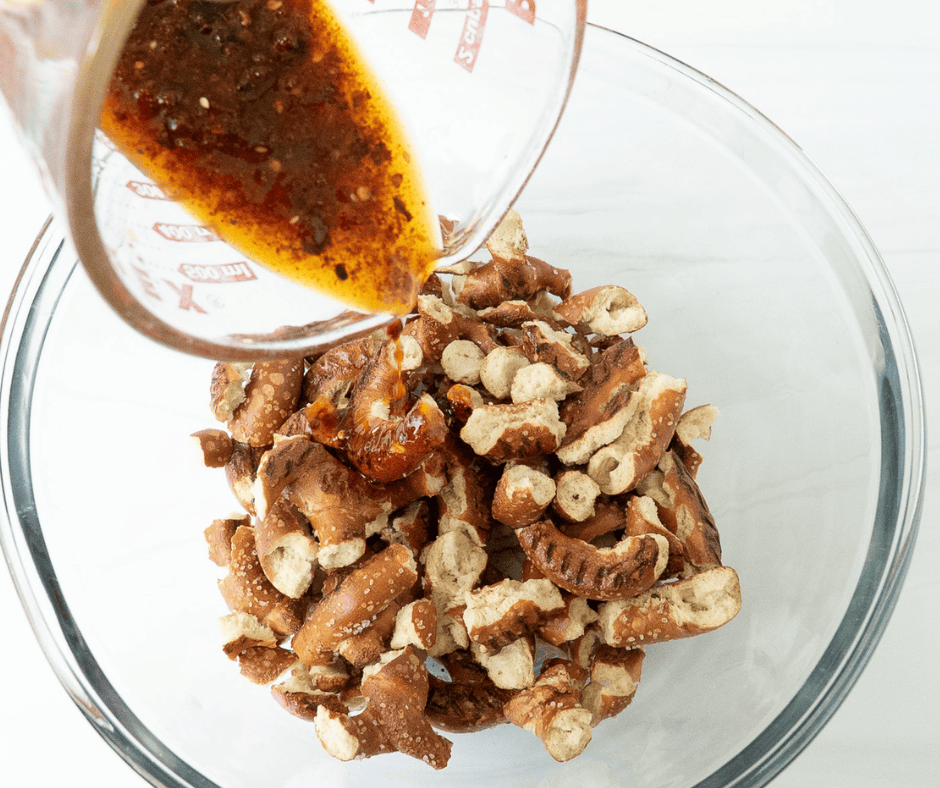 Two side-by-side process photos: the left shows a rich red chili glaze being poured over broken sourdough pretzels in a bowl; the right shows a hand adding mixed sesame seeds to the coated pretzels.