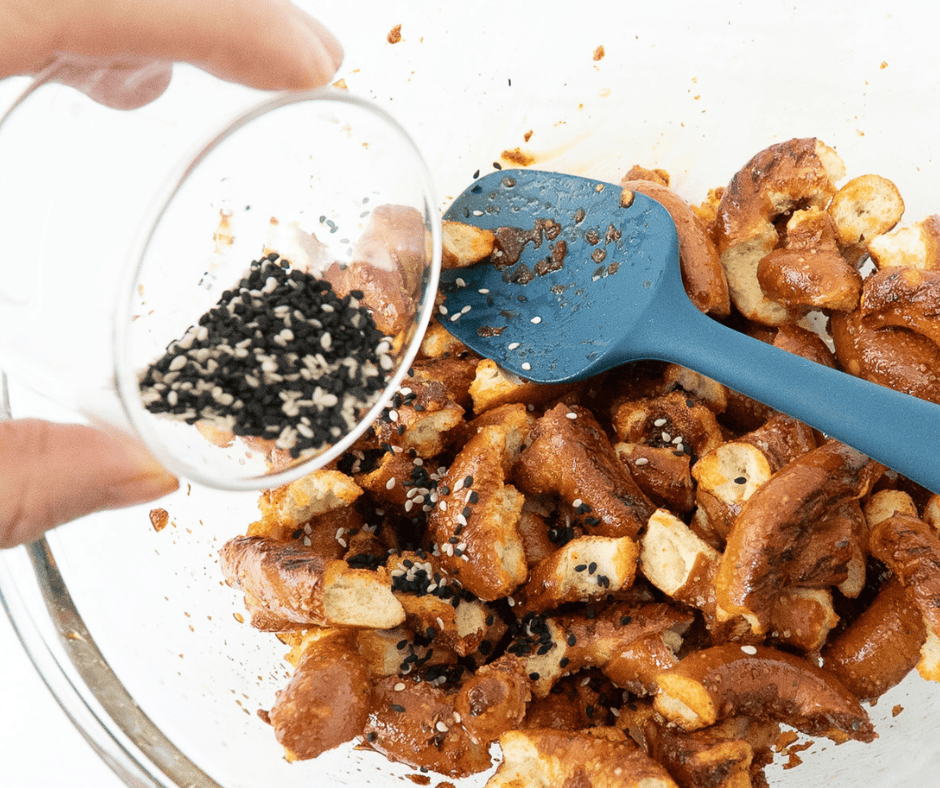 Two side-by-side process photos: the left shows a rich red chili glaze being poured over broken sourdough pretzels in a bowl; the right shows a hand adding mixed sesame seeds to the coated pretzels.