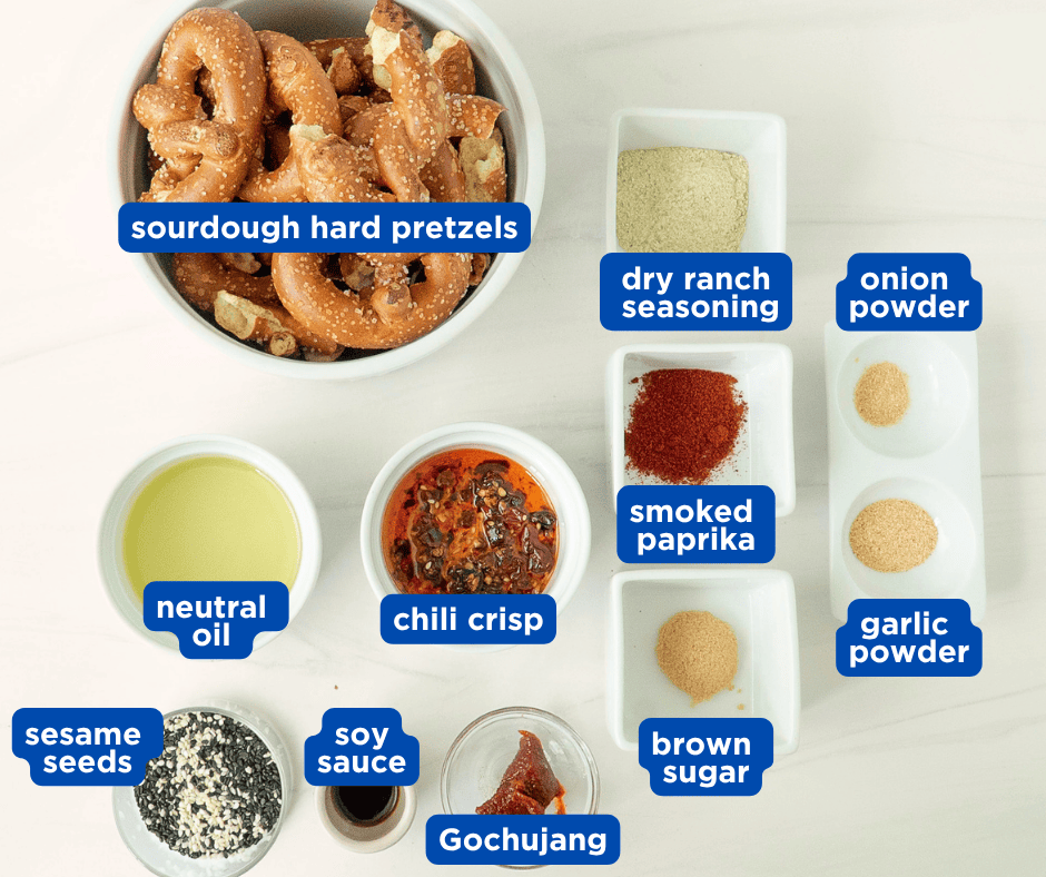 A top-down food styling layout showing bowls of ingredients for seasoned pretzels including sourdough pretzels, neutral oil, chili crisp, dry ranch dressing powder, smoked paprika, garlic powder, onion powder, soy sauce, sesame seeds, brown sugar, and a tub of gochujang.