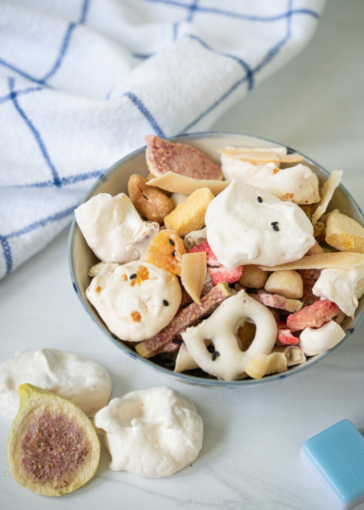 Crunchy meringue snack mix in a blue-rimmed bowl with yogurt pretzels, freeze-dried fruit, coconut chips, and cashews on a striped linen