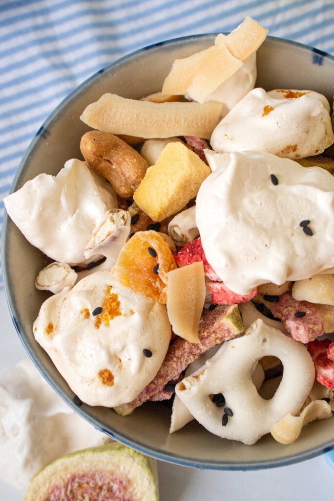 Close up of meringue snack mix in a blue bowl with freeze-dried fruit, nuts, and yogurt pretzels, with a fresh fig and mahjong tile alongside